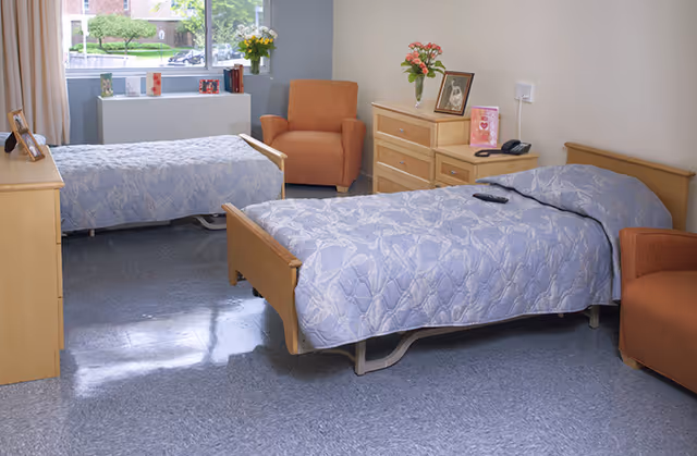 A bright, clean room with two single beds covered in light blue patterned bedspreads. Each bed has a wooden headboard and footboard. There are two orange armchairs, one near the window and one near the bed in the foreground. A wooden dresser with flowers, framed photos, and a telephone is against the wall. A large window lets in natural light and shows greenery outside.