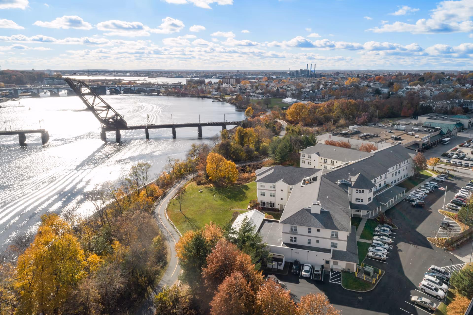 Aerial view of a multi-wing senior living building and parking lot beside a river with a bridge and autumn trees.