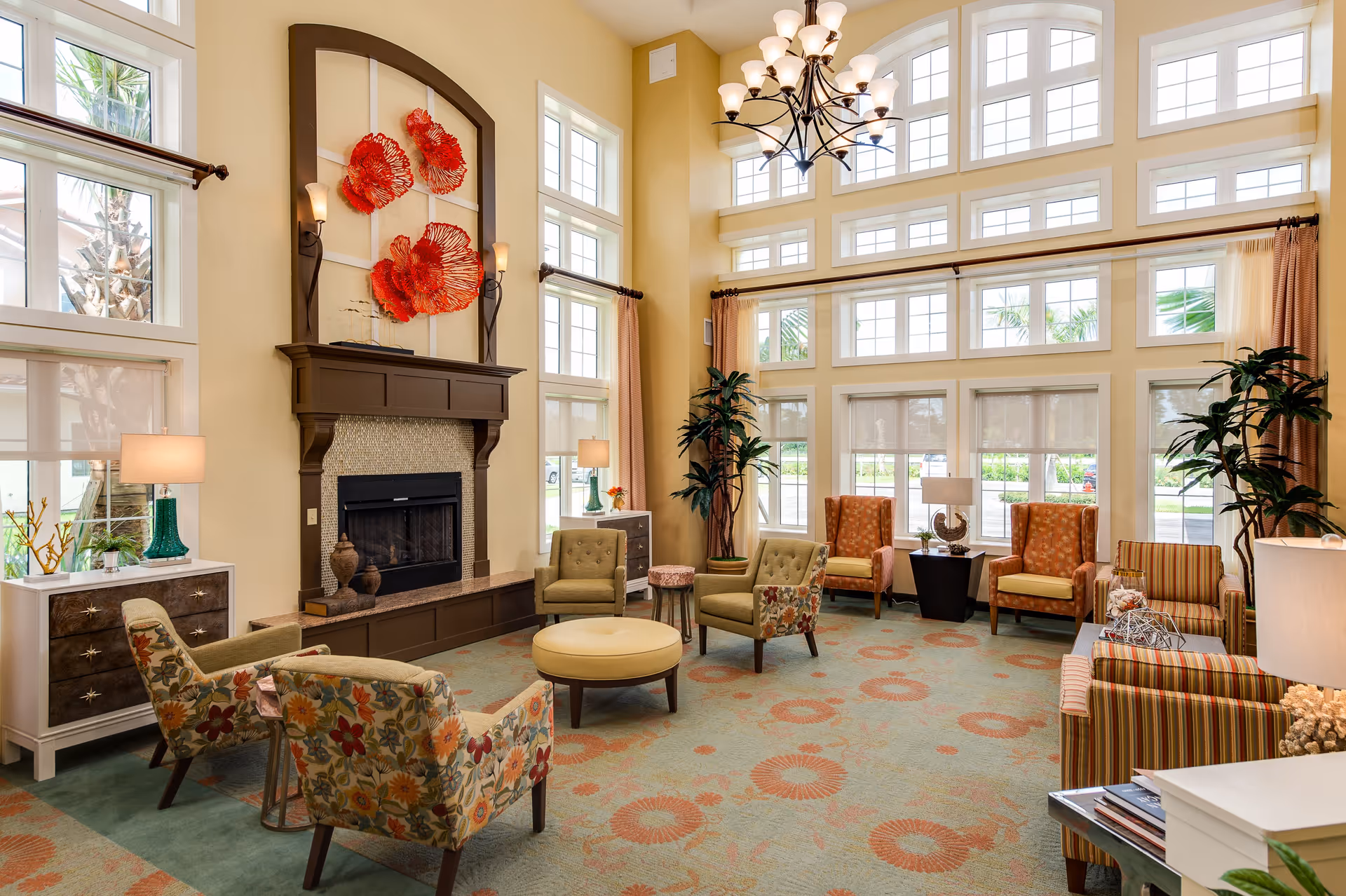 A spacious and well-lit living room with large windows and high ceilings. The room features a fireplace with decorative red wall art above it, multiple armchairs with floral and striped patterns, a round ottoman in the center, side tables with lamps, and tall indoor plants. The carpet has a floral pattern in soft colors, and a chandelier hangs from the ceiling.