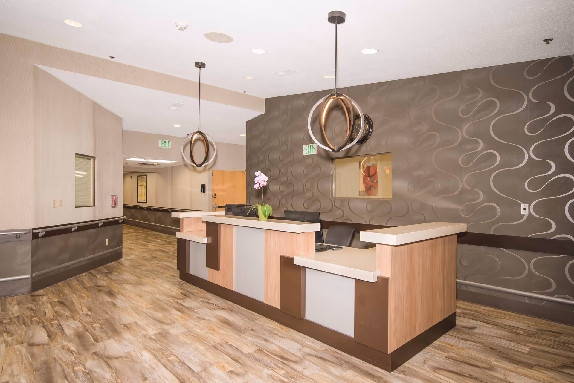 Reception area inside Aperion Care Tolleston Park featuring a modern front desk with a light wood finish and beige countertops. Two contemporary pendant lights hang from the ceiling above the desk. The wall behind the desk has a textured, wavy pattern in brown tones with a small window opening. The floor has a wood-like finish, and there is a hallway extending to the left with handrails along the walls.