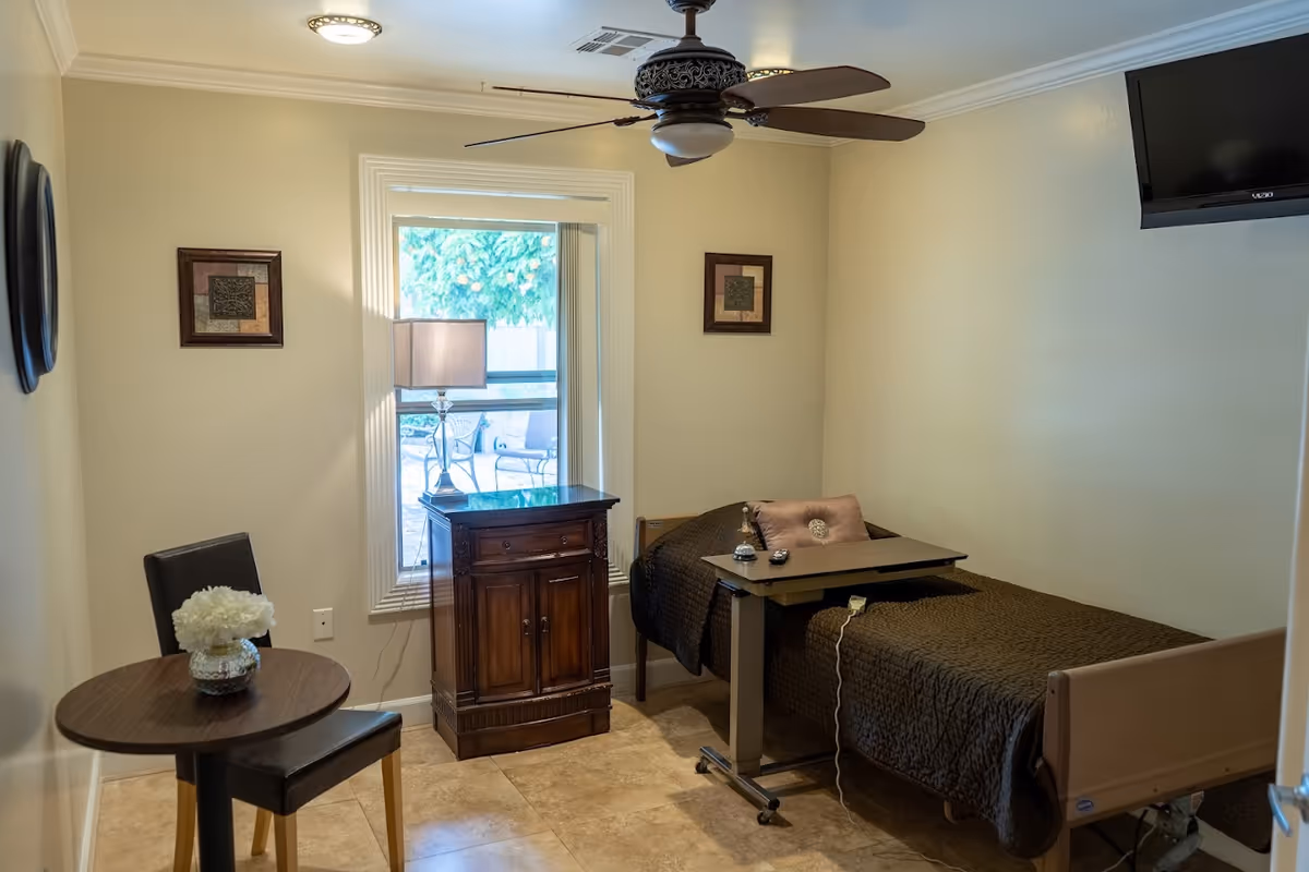 Small private bedroom with a single hospital-style bed, overbed table, wooden cabinet, small round table and chair, ceiling fan and wall-mounted TV.