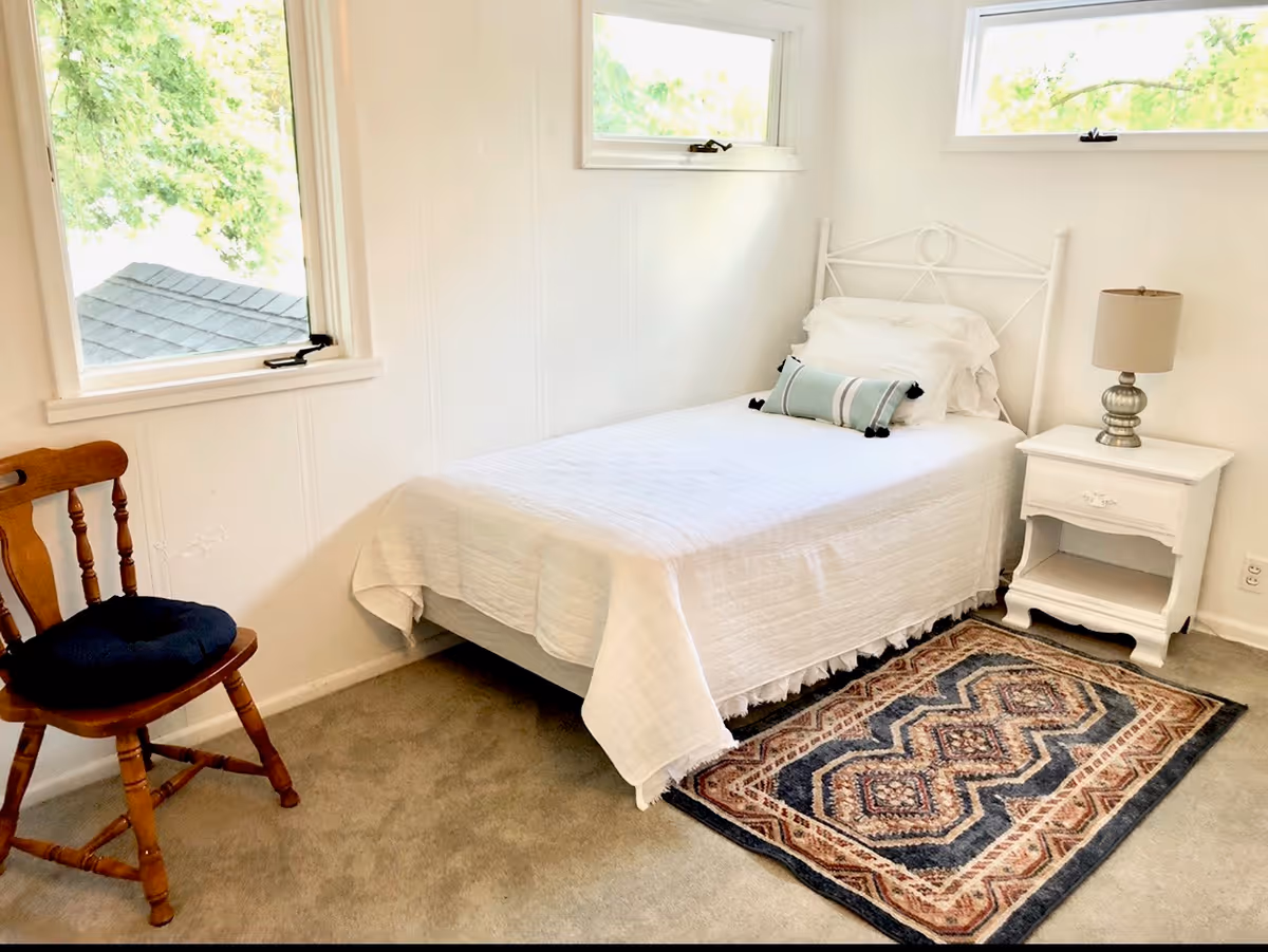 A small, bright bedroom with a single bed covered in white bedding and a decorative pillow. Next to the bed is a white nightstand with a silver lamp. A wooden chair with a dark cushion is placed near a window showing green foliage outside. A patterned rug lies on the carpeted floor beside the bed.