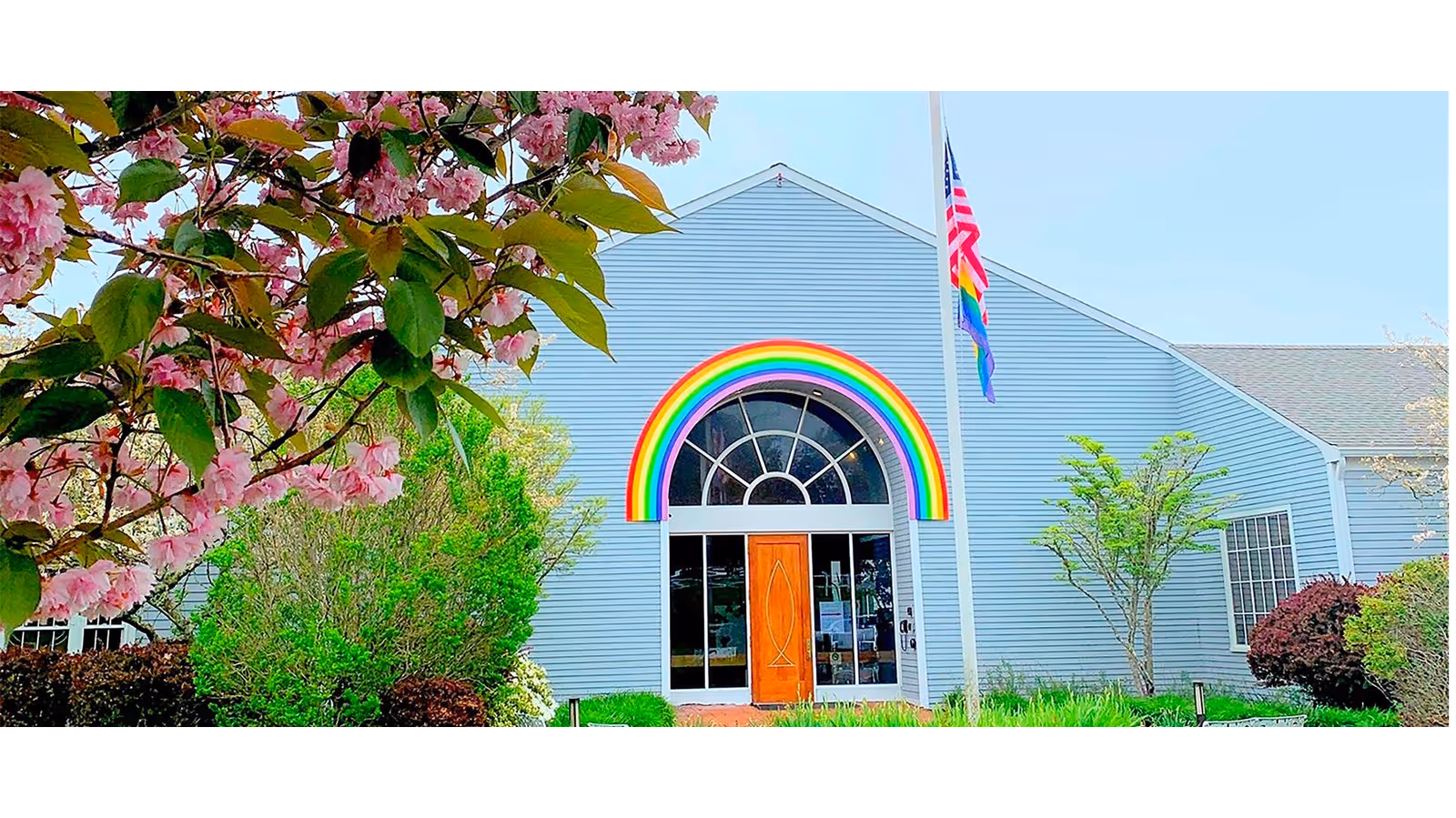 Front exterior view of a light blue building with a large arched window above a wooden door. A rainbow decoration arches over the window. There is a flagpole with an American flag and a rainbow flag in front of the building. Pink flowering tree branches and green bushes frame the scene.