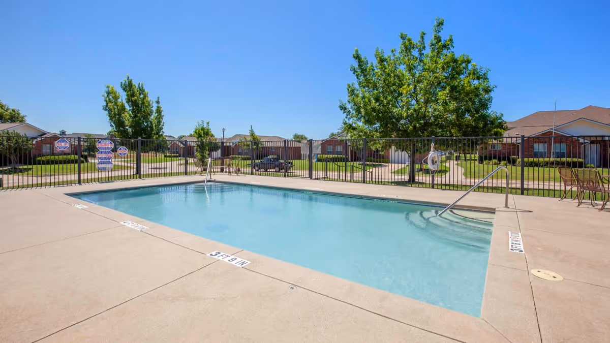 Outdoor swimming pool with clear blue water surrounded by a concrete deck and black metal fence. There are trees and residential-style buildings in the background under a clear blue sky.
