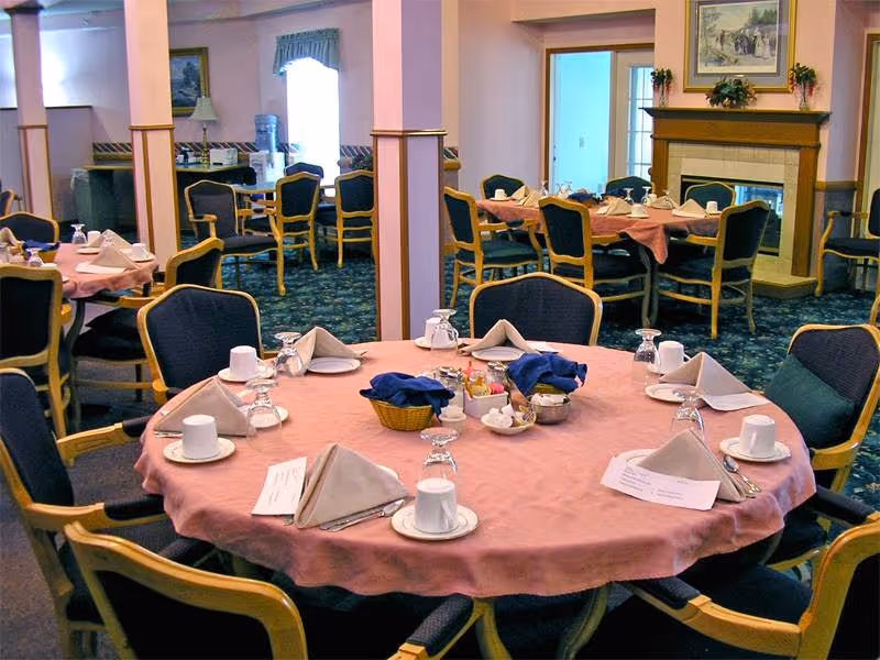 Dining room with round tables set for meals, pink tablecloths, folded napkins, cups, and cushioned chairs.