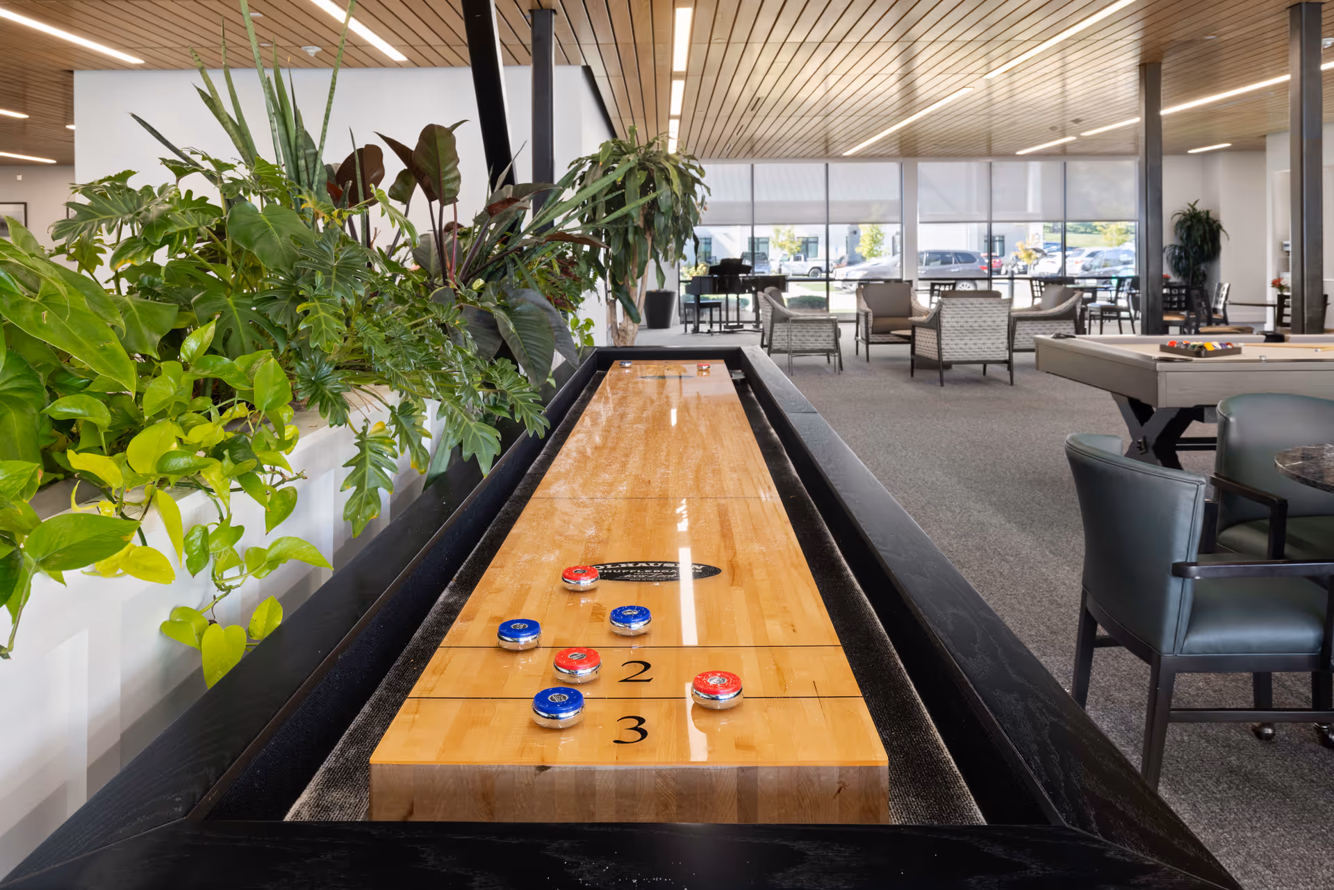 Indoor recreational area in a senior living facility featuring a shuffleboard table with red and blue pucks, surrounded by green plants on the left. In the background, there are several seating areas with chairs and tables, large windows letting in natural light, and a pool table on the right side.