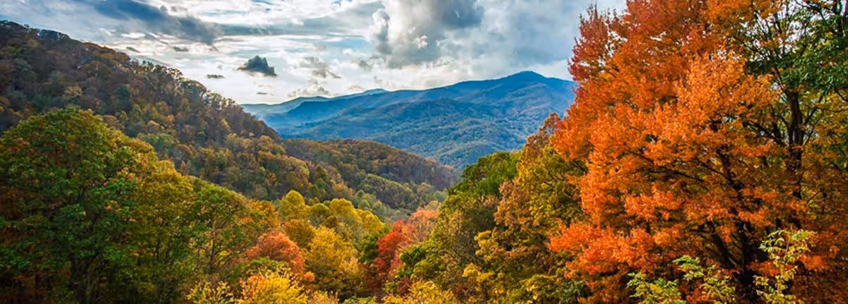 A scenic view of a mountainous landscape with dense forest in autumn colors, including green, yellow, orange, and red foliage under a partly cloudy sky.