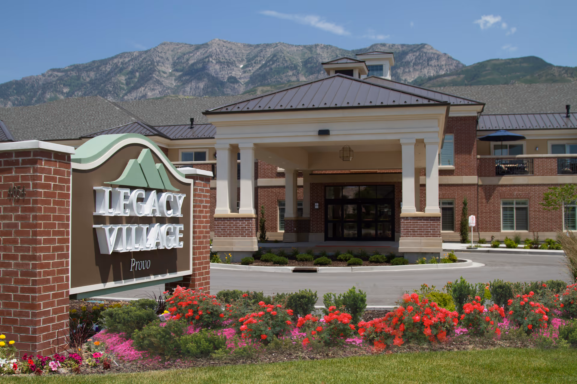 Entrance of Legacy Village of Provo senior living facility with a large sign in front, landscaped flower beds with red and pink flowers, and a building with a covered driveway and mountains in the background under a clear blue sky.