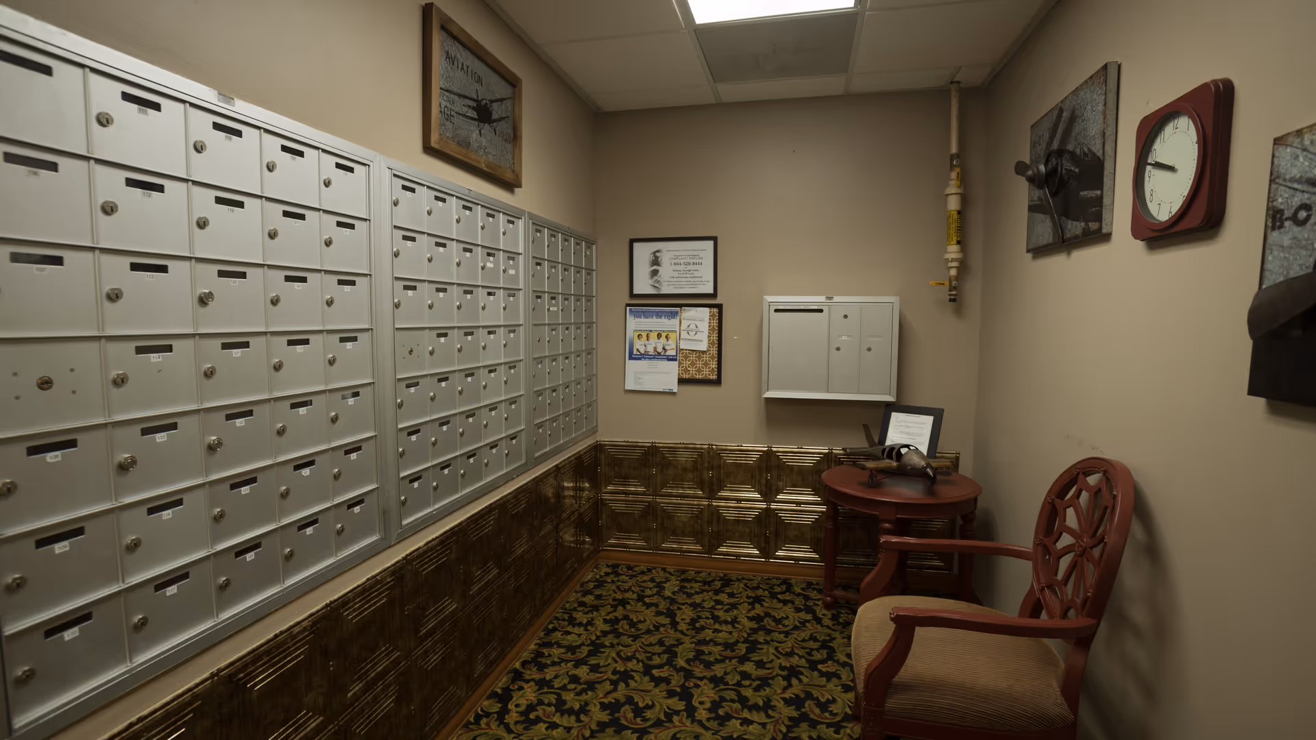 A small interior mailroom with rows of wall-mounted mailboxes, a patterned carpet, and a chair and side table under framed artwork.