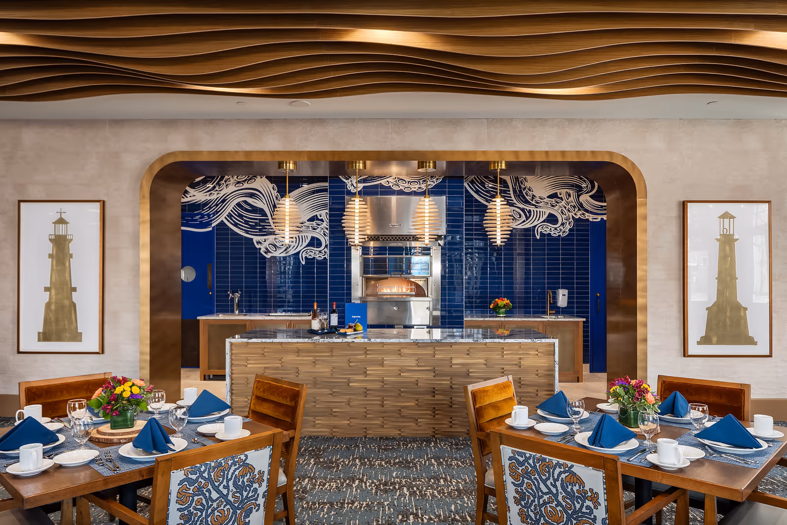 An elegant dining room with set tables and napkins in front of an open kitchen with blue tile, pendant lights, and a decorative wooden ceiling.