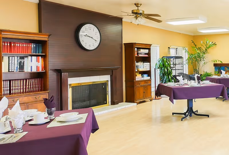 A dining room with tables covered in purple tablecloths set with white plates, cups, and folded napkins. The room features a wooden fireplace with a large clock above it, two wooden bookshelves filled with books and items, several green plants, and large windows letting in natural light.