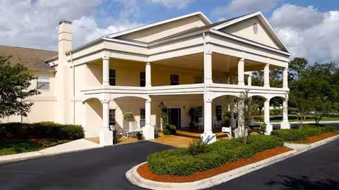 Two-story beige senior living building with a covered front entrance and landscaped circular driveway.