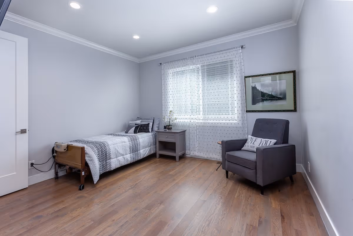 A simple bedroom with a single bed, nightstand by a curtained window, and a gray armchair on hardwood floors.