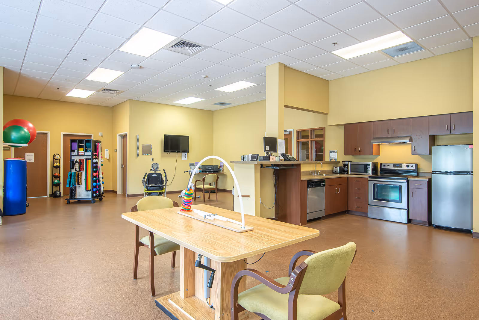 Bright communal activity and kitchenette area with a wooden table and chairs, exercise equipment and storage racks, and stainless steel appliances.