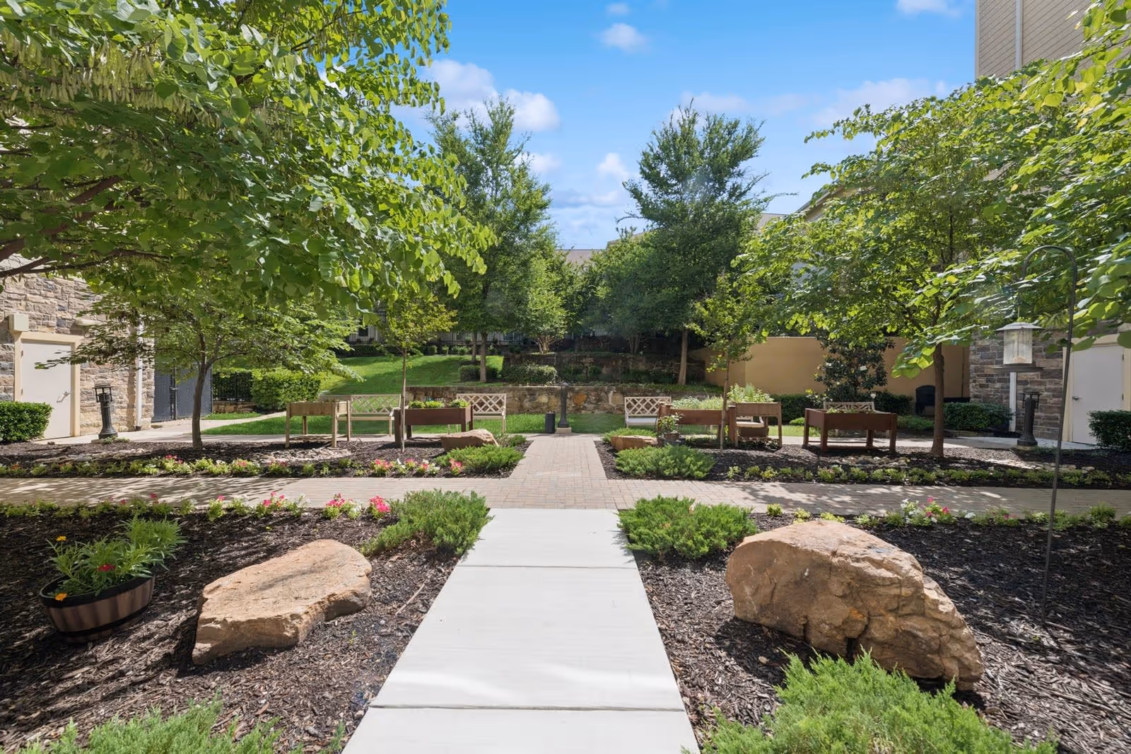 A landscaped outdoor courtyard area with a paved walkway leading to benches surrounded by trees, shrubs, and large rocks under a blue sky with some clouds.