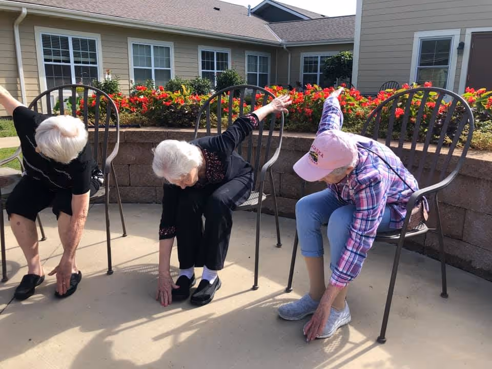 Three elderly women sitting on metal chairs outdoors in front of a building with flower beds, stretching by reaching down towards their feet with one arm extended back.