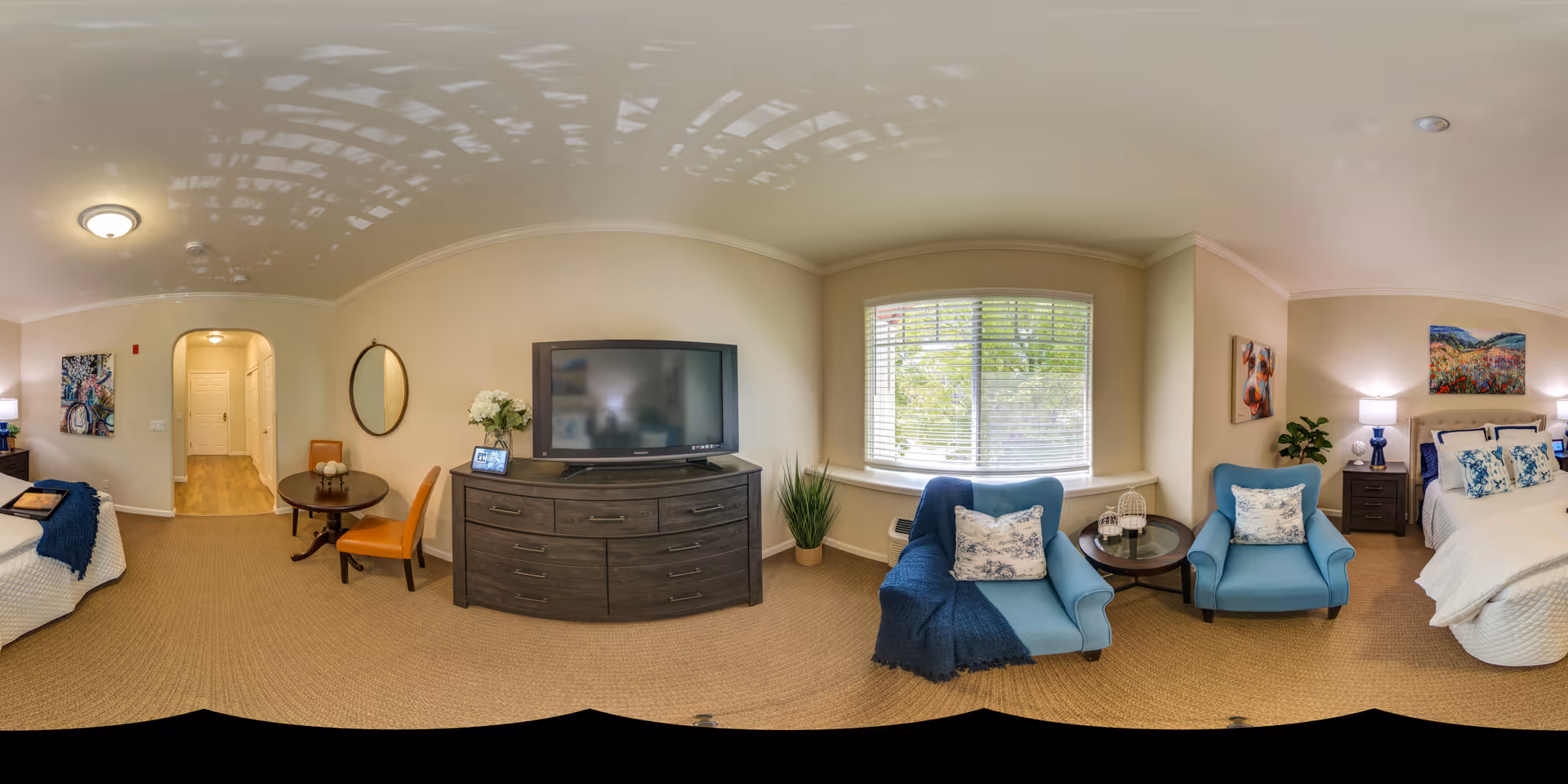 A well-lit senior living facility room with a bed on the right side, two blue armchairs with pillows near a window with blinds, a round side table between the chairs, a dark wooden dresser with a flat-screen TV on top, a small round dining table with two orange chairs, and decorative wall art and plants throughout the room.