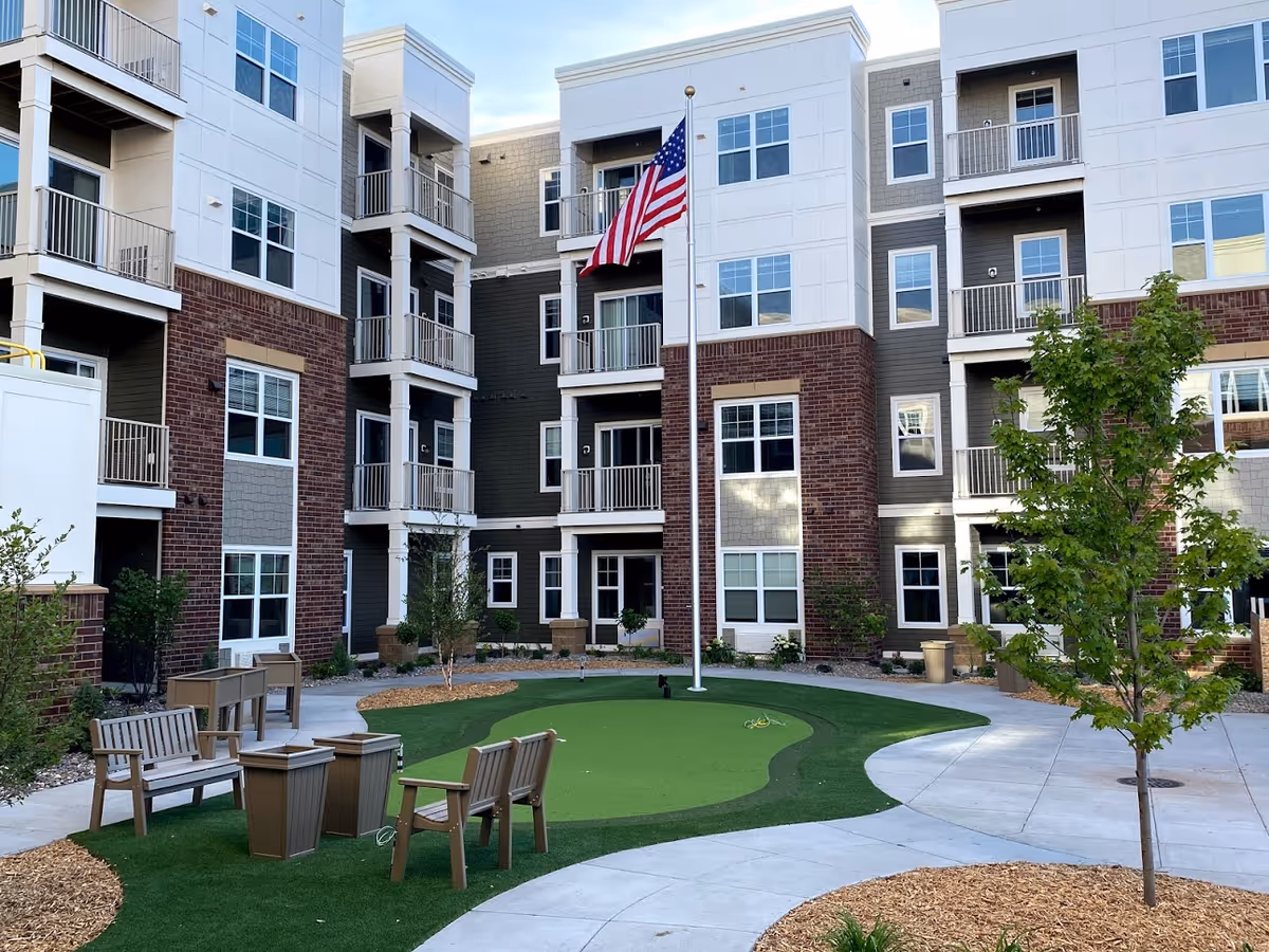 Outdoor courtyard area of a multi-story senior living facility with a putting green, several wooden benches and chairs, a flagpole with an American flag, trees, and landscaped walkways.