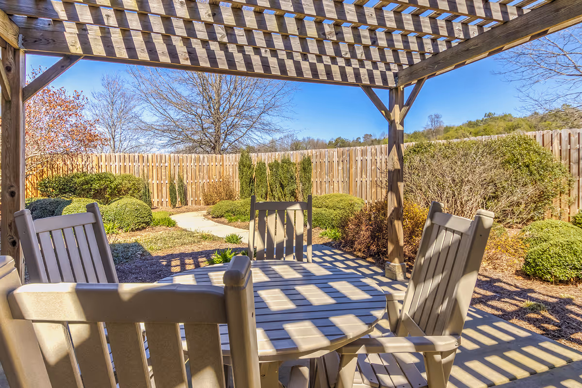 Outdoor seating area with a round table and four chairs under a wooden pergola, surrounded by bushes and trees with a wooden fence in the background under a clear blue sky.
