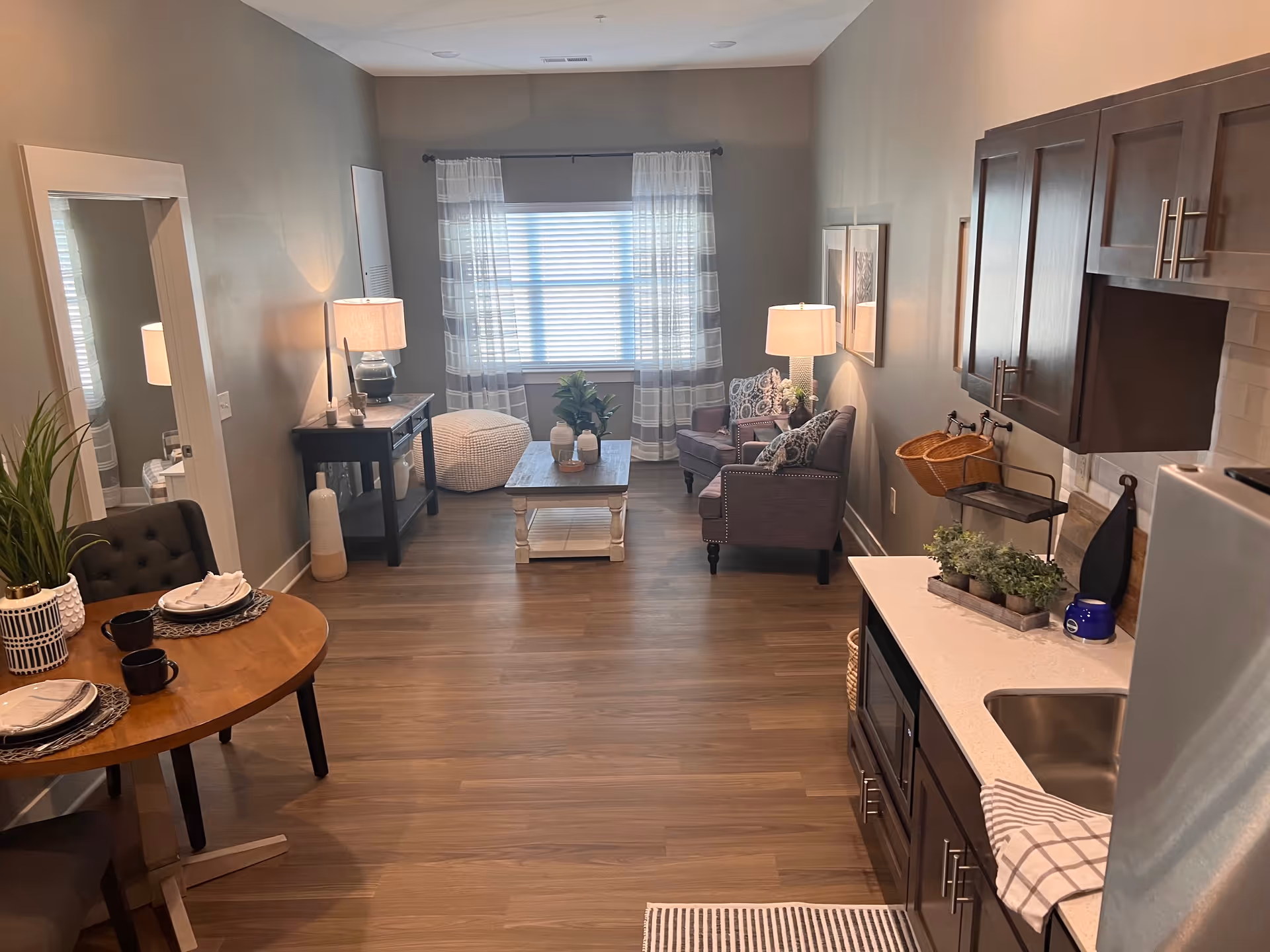 A cozy living space with a small round dining table set for two on the left, a large mirror on the wall, and a kitchen area with dark cabinets and a white countertop on the right. The room has wooden flooring, a coffee table in the center, two armchairs with patterned cushions, a pouf near the window with sheer curtains, and two table lamps providing warm lighting.