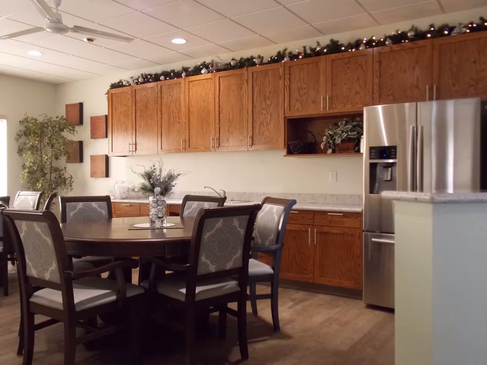 Dining area with a round table and upholstered chairs in front of wood kitchen cabinets and a stainless steel refrigerator.