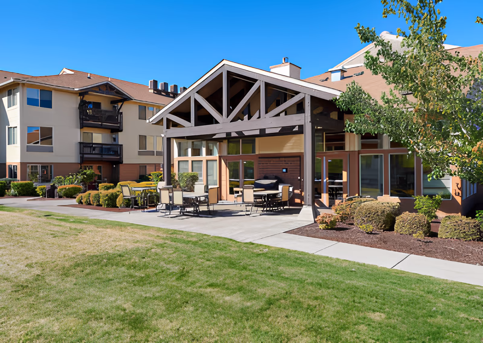 Outdoor patio area at Elliott Residence with tables and chairs under a wooden pergola, surrounded by bushes and trees, adjacent to a multi-story residential building under a clear blue sky.