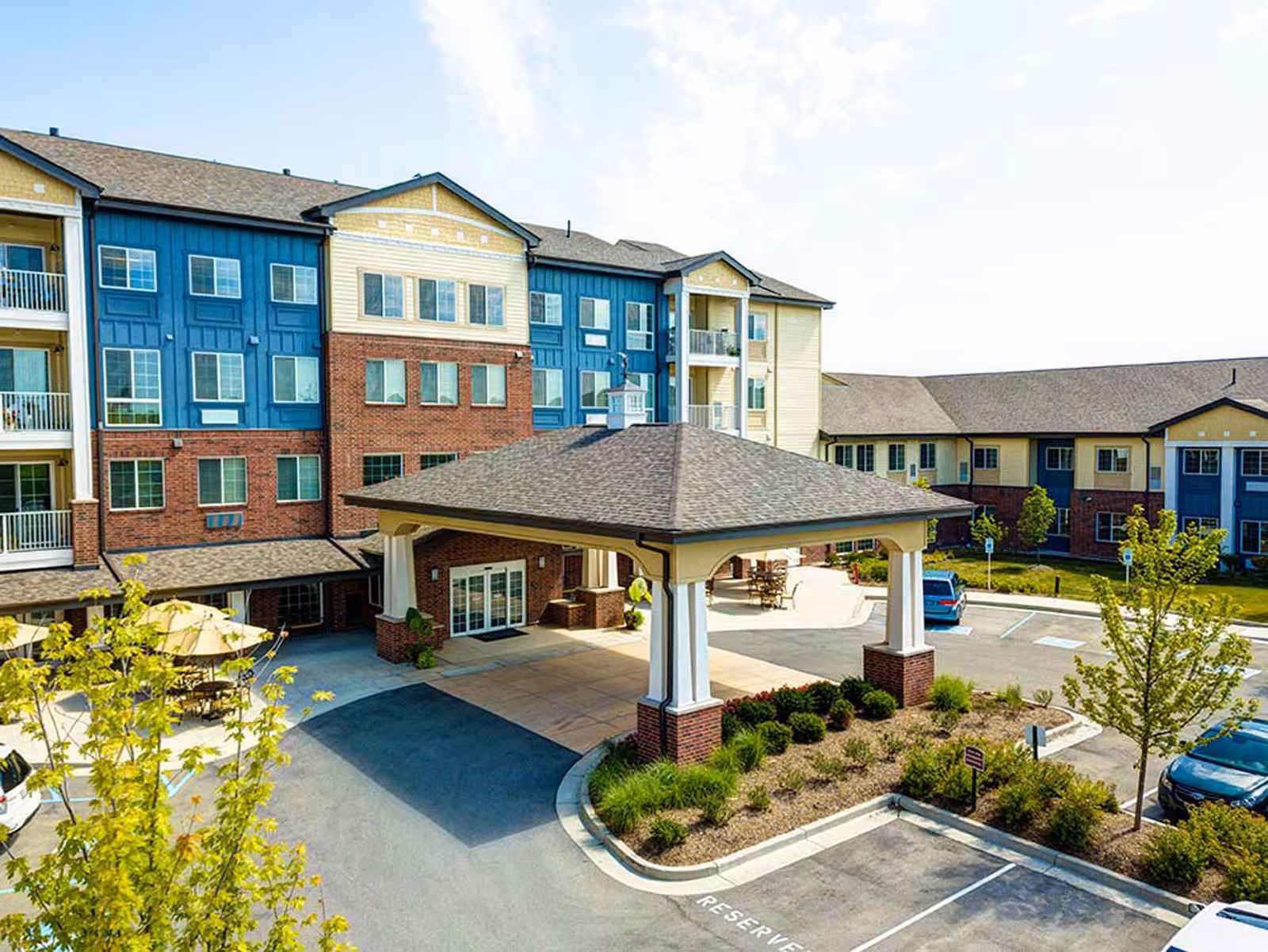 Exterior view of a multi-story senior living facility building with a covered entrance driveway, landscaped greenery, parking spaces, and outdoor seating areas with umbrellas.