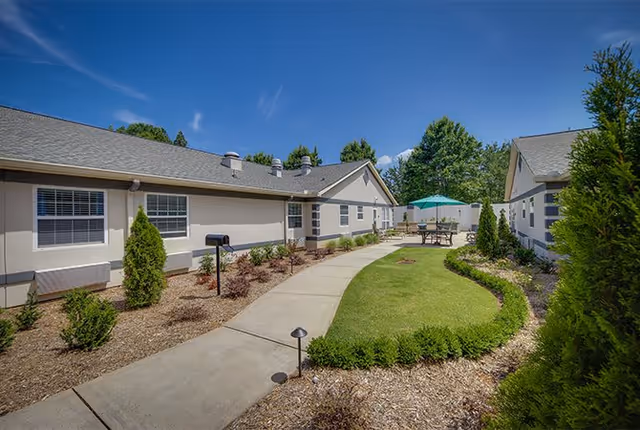 Outdoor view of a senior living facility courtyard with a concrete walkway, green lawn, small shrubs, and two single-story buildings on either side under a clear blue sky. There is a patio table with an umbrella in the background.