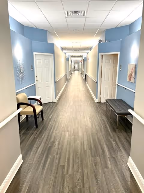 A long, well-lit hallway in an assisted living facility with light blue and beige walls, wood-patterned flooring, white doors on both sides, handrails along the walls, a beige chair on the left, and a black bench on the right.