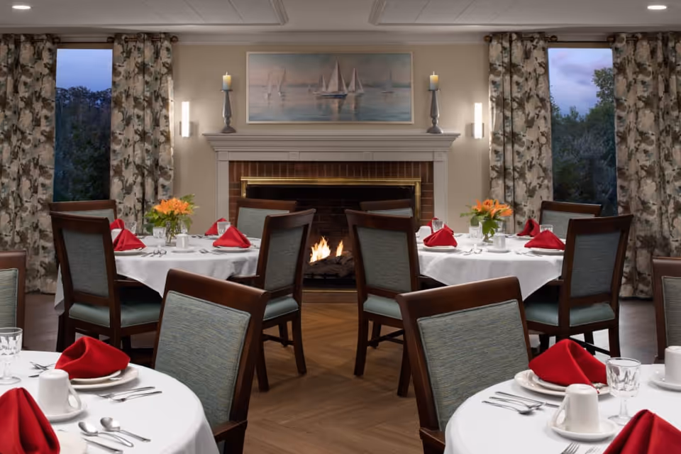 A cozy dining room with round tables set with white linens and red napkins, wooden chairs, floral centerpieces, and a lit fireplace beneath a sailboat painting.