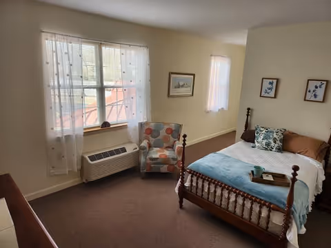A cozy bedroom with a wooden bed featuring a white bedspread, blue throw blanket, and decorative pillows. There is a small wooden tray with a cup on the bed. Next to the bed is a colorful patterned armchair. The room has beige walls with framed artwork and two windows with sheer white curtains letting in natural light. A heating/cooling unit is beneath one window.