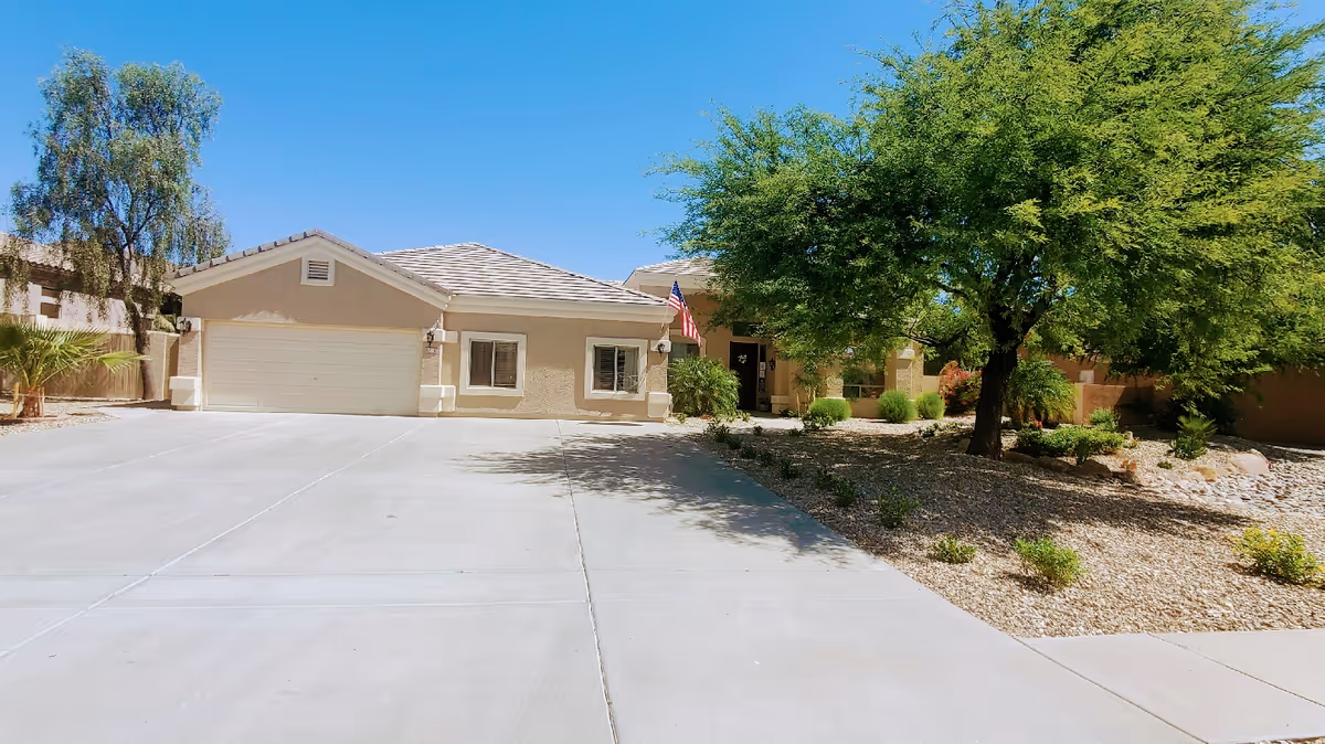 Front exterior view of a single-story residential building with a two-car garage, beige stucco walls, a tiled roof, and an American flag near the entrance. The driveway is wide and paved, and there is a landscaped area with trees and shrubs on the right side under a clear blue sky.