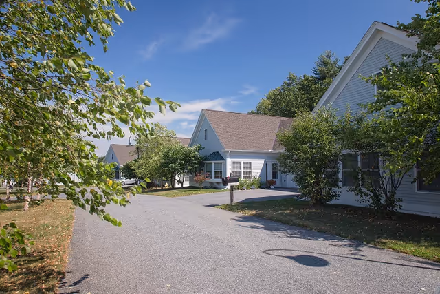 A paved driveway lined with trees and shrubs leads to several single-story residential buildings with white siding and brown roofs under a clear blue sky.