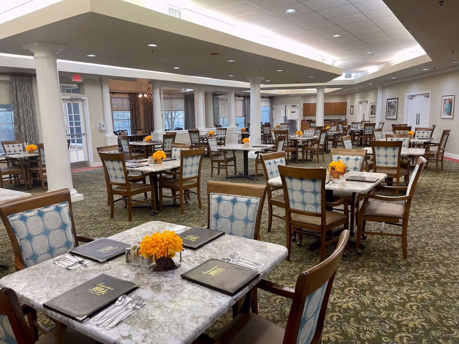 A spacious dining room in an assisted living facility with multiple tables and chairs arranged neatly. Each table has a small orange flower centerpiece and menus labeled 'Independence Hill'. The room features carpeted floors, white columns, large windows with curtains, and bright overhead lighting.
