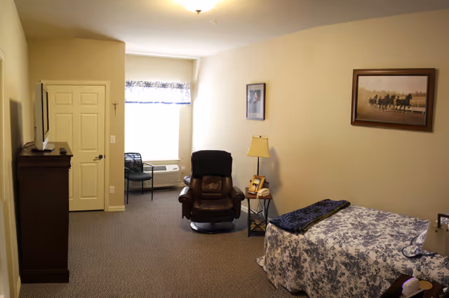 A small, cozy bedroom with beige walls and carpeted floor. The room features a single bed with a floral bedspread, a brown leather recliner chair, a small side table with a lamp and picture frames, a window with a valance, a wall-mounted TV, and framed pictures on the walls.