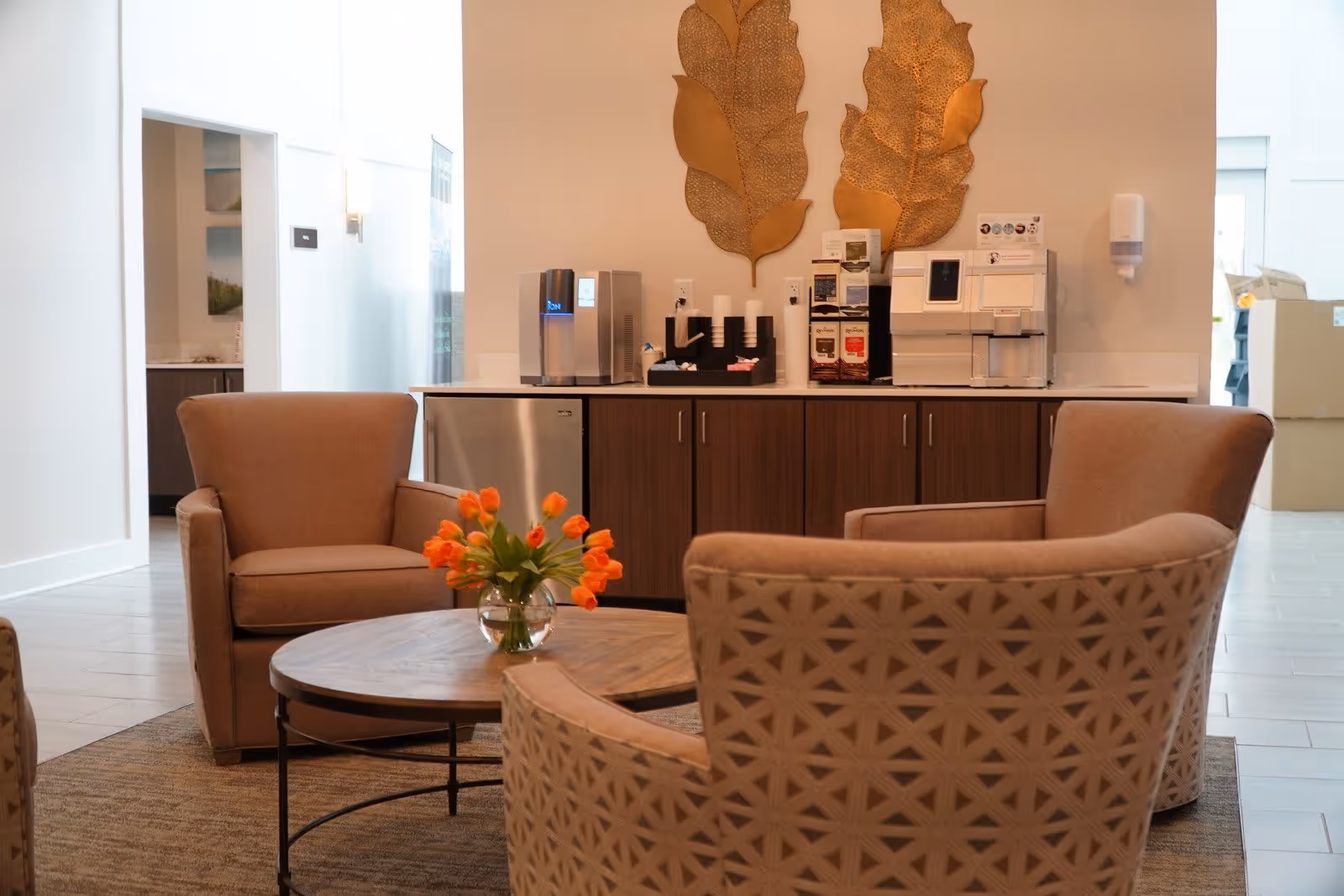 Cozy seating area with upholstered armchairs around a round coffee table holding a vase of orange tulips and a coffee station along the back wall.