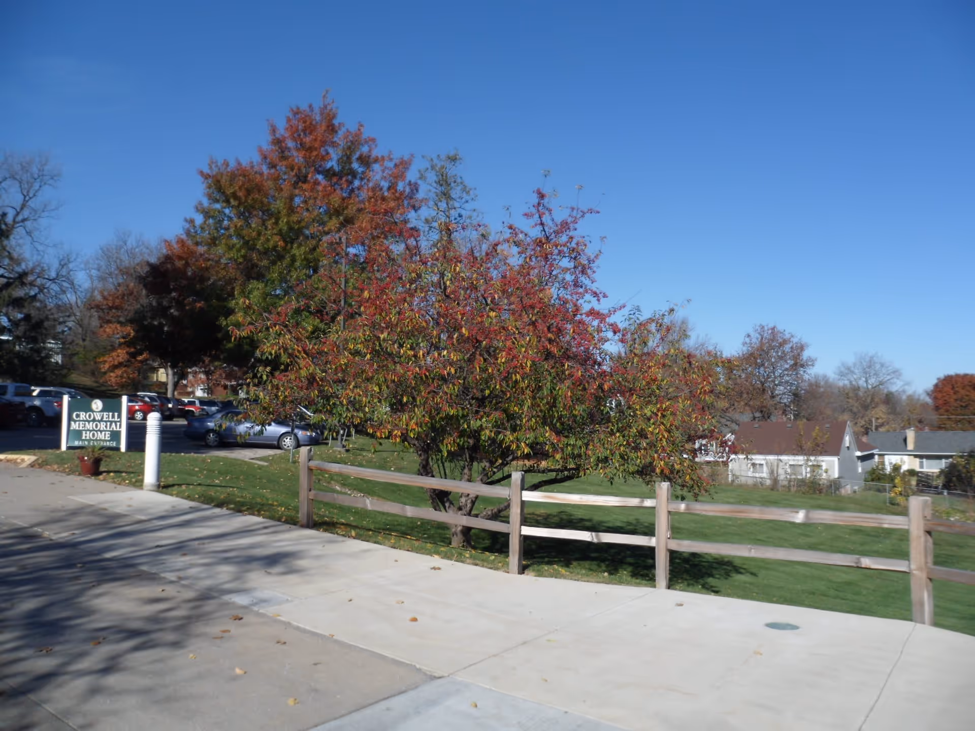 Outdoor view of Crowell Memorial Home showing a parking lot with several cars, a green sign with white text indicating the main entrance, a wooden fence, and trees with autumn foliage under a clear blue sky.