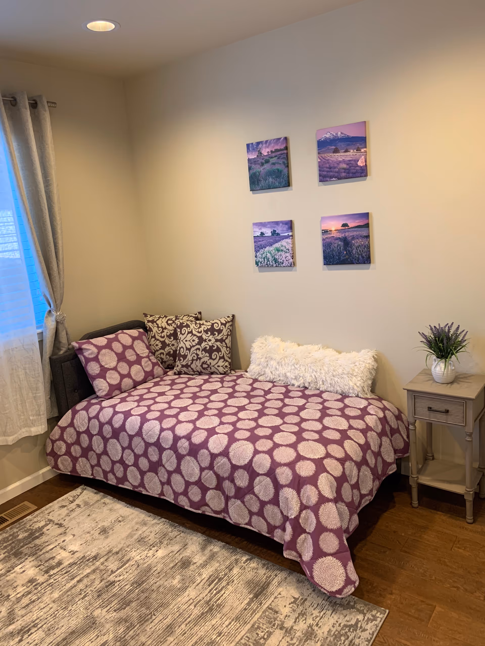 A cozy bedroom with a single bed dressed in purple patterned bedding, decorative pillows, a small nightstand with a plant, and four landscape pictures on the wall.