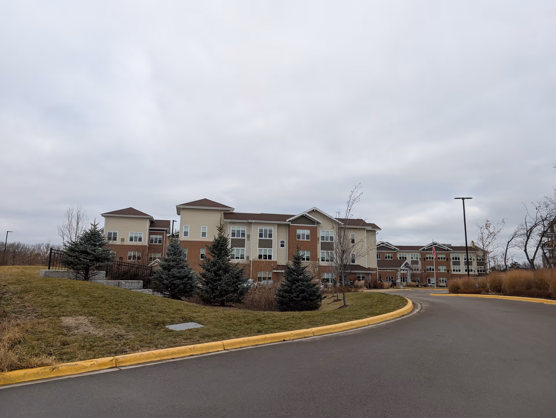 Exterior view of a multi-story senior living building with a curved driveway, grass and evergreen trees under a cloudy sky.