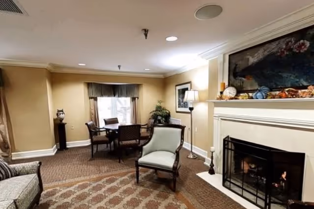 Cozy common room with upholstered chairs, a small dining table by a window, and a lit fireplace with mantel decor.