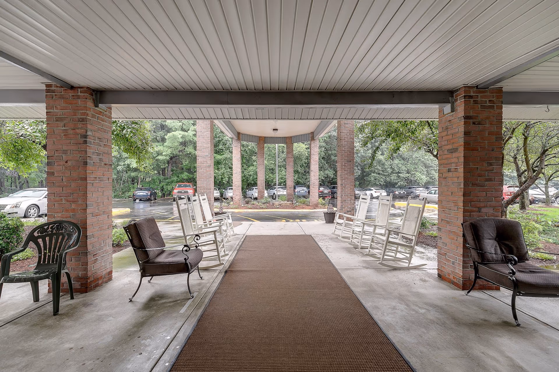Covered outdoor seating area at the entrance of a senior living community with multiple rocking chairs and cushioned chairs arranged along the sides. The area is supported by brick columns and overlooks a parking lot with trees and greenery in the background.