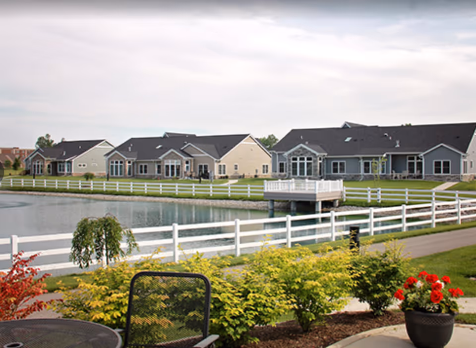 View of a peaceful senior living community with single-story houses surrounding a pond. There is a white fence along the pond, a small dock extending over the water, and well-maintained landscaping with bushes and flowers in the foreground. A black metal chair and table are partially visible on a patio area.