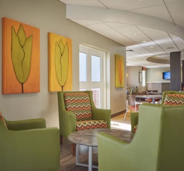 A cozy seating area in a senior living facility with green armchairs featuring colorful zigzag patterned cushions arranged around a round wooden table. The walls are decorated with three paintings of stylized green leaves on orange and yellow backgrounds. In the background, there are more tables and chairs, a TV mounted on the wall, and a ceiling with recessed lighting.