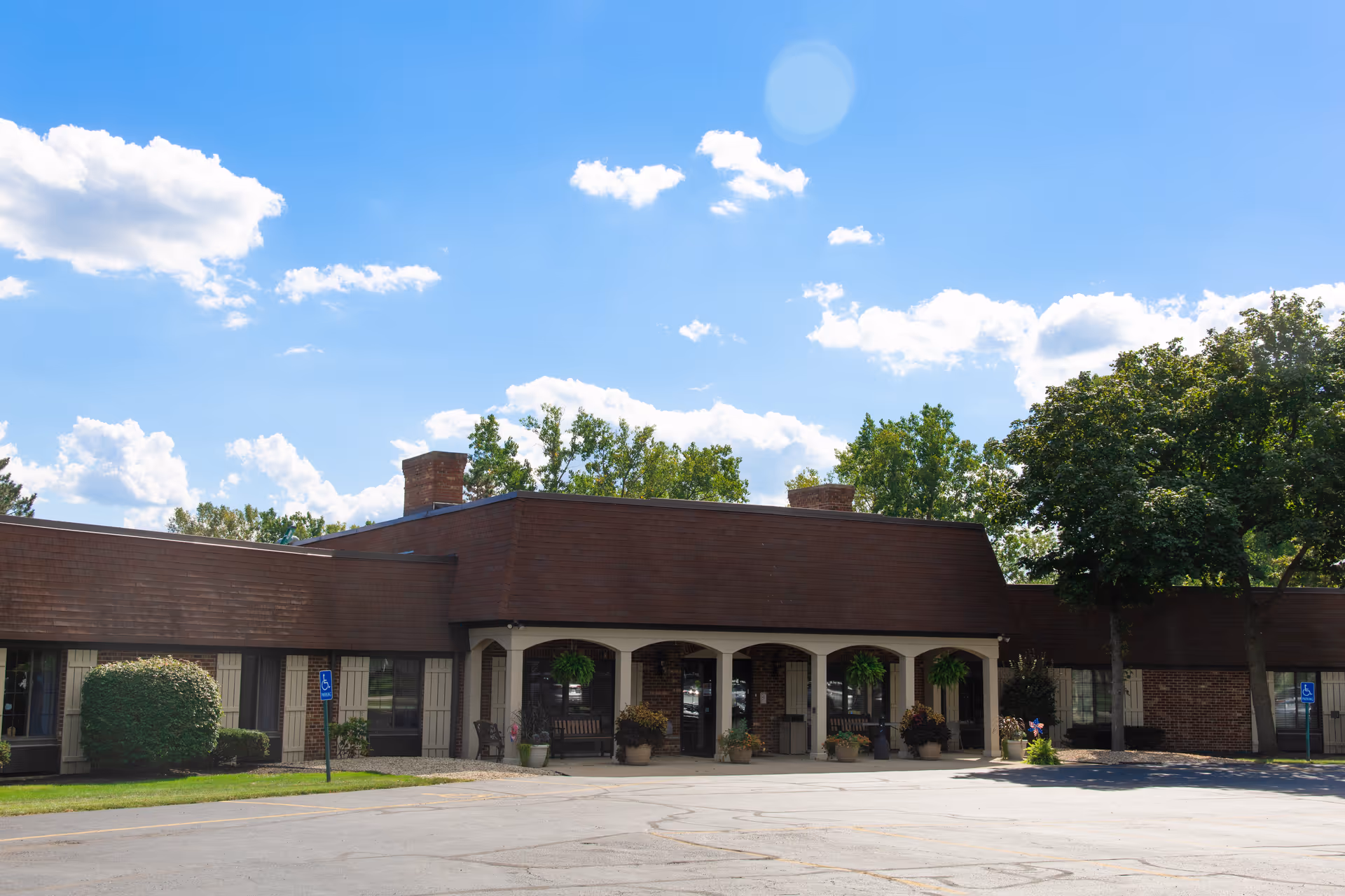 Front exterior of a single-story brick senior living building with an arched covered entrance, hanging plants, and an empty parking lot under a blue sky.