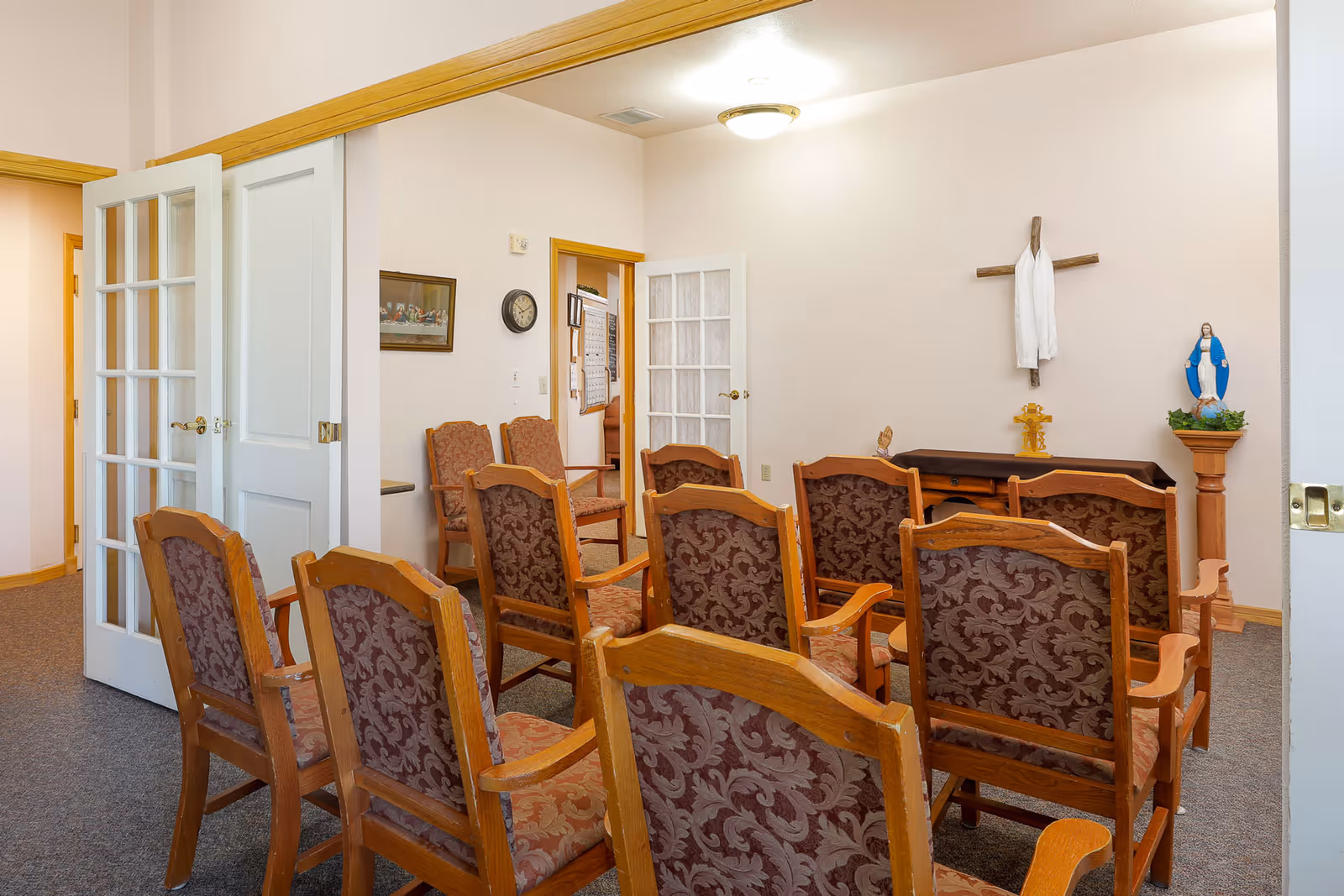 A small chapel or prayer room with wooden chairs arranged in rows facing a wall with a cross draped with a white cloth, a small altar table, and a statue of the Virgin Mary on a pedestal. The room has light-colored walls, carpeted floor, and a ceiling light fixture. There are double doors with glass panes on the left side.