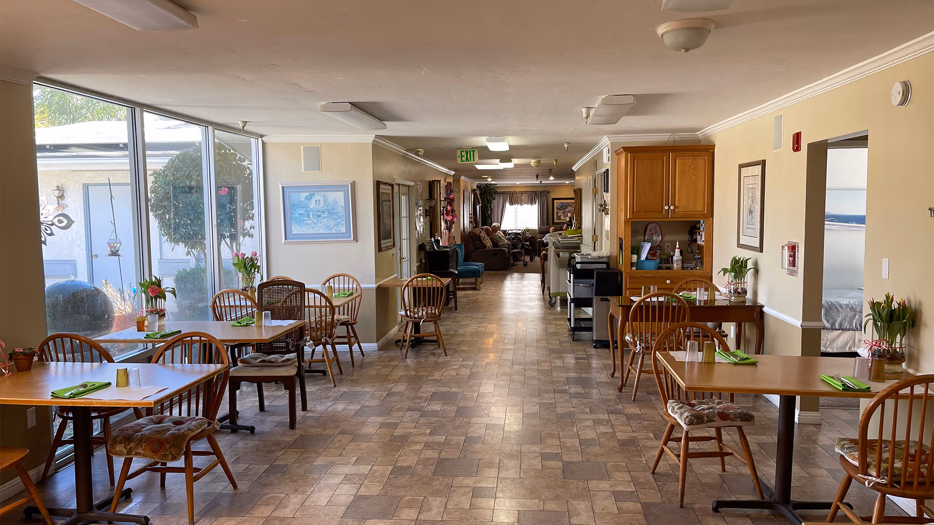 Long dining and common area in an elderly care facility with tables and chairs along a windowed wall and a seating area down the corridor.