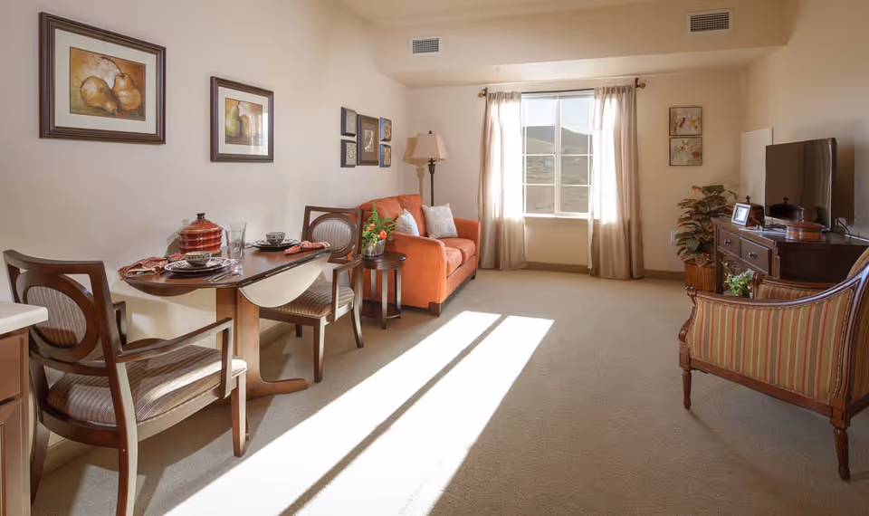 A bright and cozy living room in a senior living facility featuring a small dining table with two chairs, an orange sofa with cushions, a side table with a flower vase, a large window with curtains letting in sunlight, a wooden TV stand with a flat-screen TV, and a striped armchair. The walls are decorated with framed artwork.