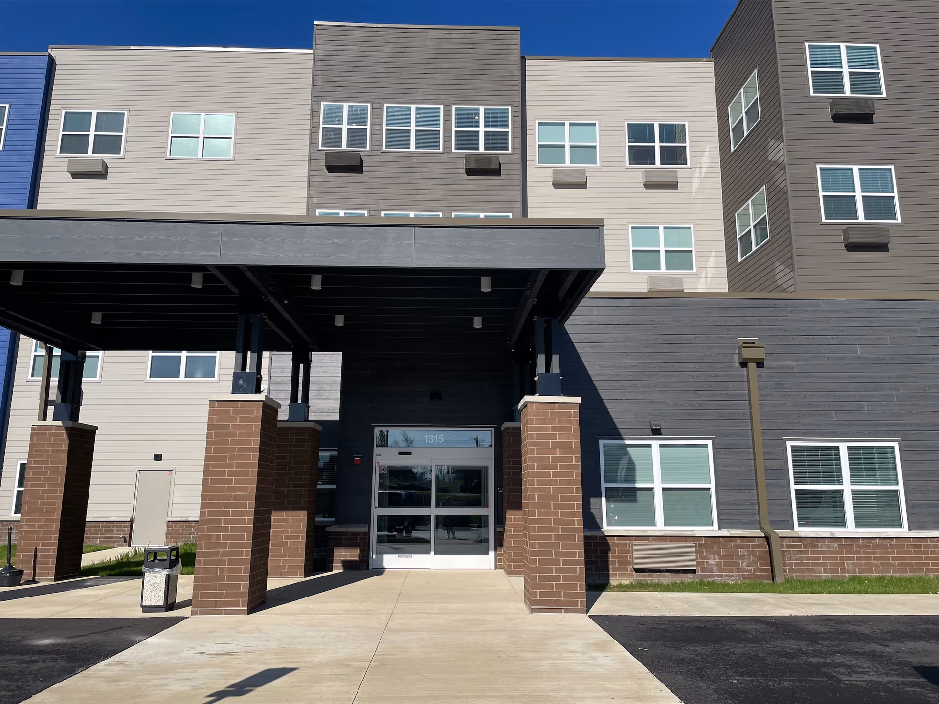 Front entrance of a multi-story modern residential building with a covered drop-off area supported by brick pillars.