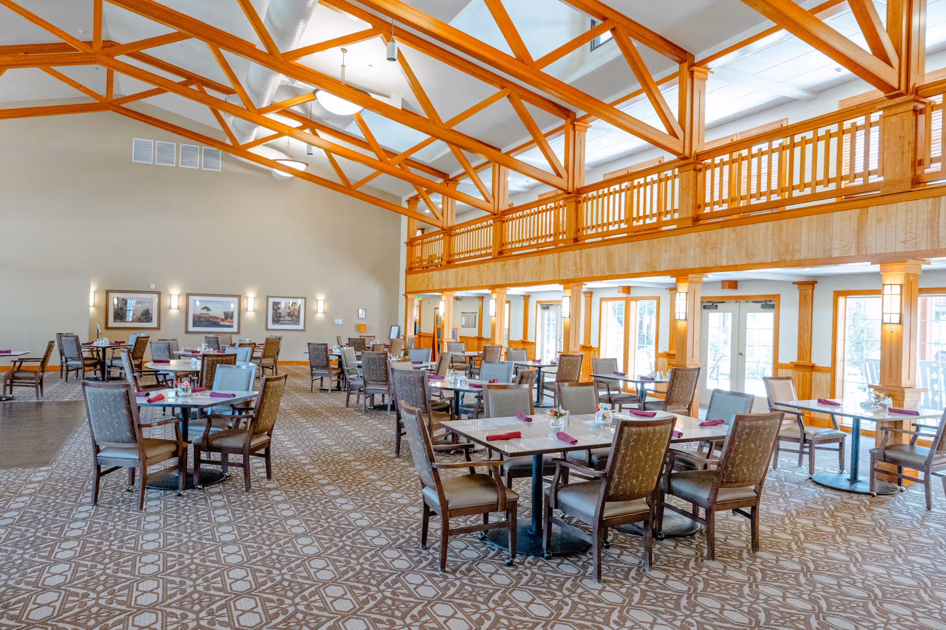 A spacious dining room with multiple tables and chairs arranged neatly. The room features a high ceiling with exposed wooden beams and a mezzanine balcony. Large windows and glass doors allow natural light to fill the space. Each table is set with napkins and small floral centerpieces.