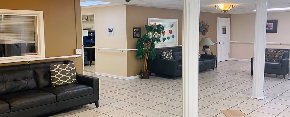 Interior view of a senior living facility common area with beige walls, tiled floor, and several black leather sofas with patterned cushions. There are white columns, a potted plant, framed artwork on the walls, and a ceiling light fixture. A window and door are visible in the background.
